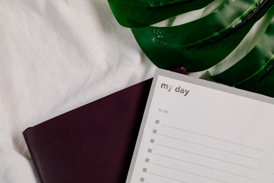 A close-up of a to-do list notebook with lush green leaves on a white textile surface.